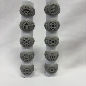 May include: A selection of grey rings, each with a distinct symbol design. The rings are presented on white cylindrical stands. The symbols include a sunburst, a lightning bolt, and geometric shapes.