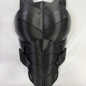 Może przedstawiać: Czarna, formowana zbroja na klatkę piersiową z symbolem nietoperza na piersi. Zbroja ma szczegółowy design z segmentowanymi płytami i wysokim kołnierzem. Ogólny kształt sugeruje kostium superbohatera, prawdopodobnie do cosplayu lub ekspozycji.