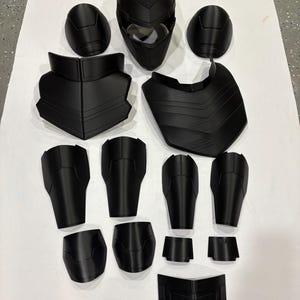 Peut inclure: Ensemble d'armure noire comprenant un casque, des protège-épaules, des plaques de poitrine et des protections pour les membres. Les composants sont disposés sur une surface blanche, présentant un ensemble complet.