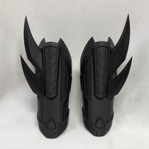 Könnte beinhalten: Paar schwarze, futuristisch aussehende Armschützer. Jeder Schützer hat eine Reihe von abgewinkelten, klingenartigen Vorsprüngen, die sich von der Oberseite erstrecken. Die Schützer haben ein segmentiertes Design mit geometrischen Mustern und sind wahrscheinlich aus Kunststoff.