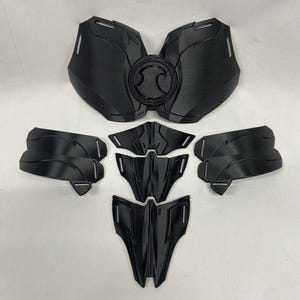 Può includere: Un set di sei pezzi di plastica nera che formano una placca per il petto per un costume. La placca per il petto ha un design circolare al centro.