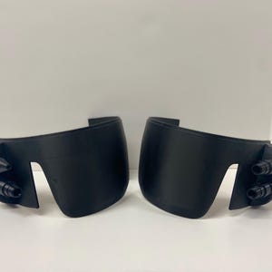 May include: Two black plastic face shields with a curved shape and multiple small holes on the sides.