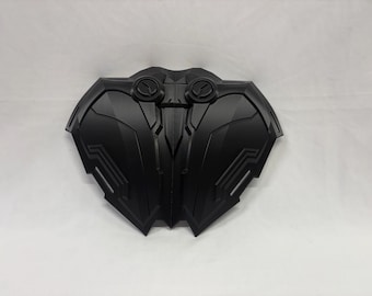 May include: A black, symmetrical shield-like object with a complex, geometric design. The object has two circular features at the top and a series of angled lines and panels. The overall shape is reminiscent of a stylized insect or mechanical component.