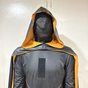 Könnte beinhalten: Ein schwarzes Kapuzenkostüm mit orangefarbenen Verzierungen. Das Kostüm hat ein strukturiertes Muster und eine vollständige Gesichtsbedeckung. Die Kapuze und der Umhang sind mit orangefarbenem Stoff gefüttert. Das Kostüm hat einen rechteckigen Aufnäher auf der Brust.