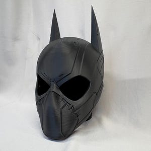 May include: A black superhero-style mask with distinctive pointed ears. The mask has eye openings, a nose and mouth section, and detailed surface paneling. It appears to be constructed from a rigid plastic or composite material.
