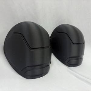 May include: Two black, helmet-shaped objects with a smooth, matte finish. Each has a distinct design with a raised section across the top and a curved lower portion. The objects are set against a plain white background.