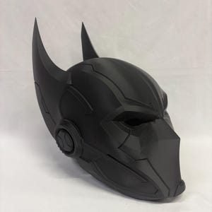 Op de afbeelding: Een matzwarte superheldenmasker met puntige oren en hoekige designelementen. Het masker heeft openingen voor de ogen en een kinbeschermer, ideaal voor cosplay of kostuums.