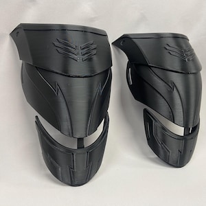Könnte beinhalten: Ein Paar schwarze, 3D-gedruckte Knieschoner mit geometrischem Design. Die Schoner haben ein erhabenes, strukturiertes Muster und ein kleines, erhabenes Symbol oben.