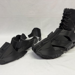 Può includere: Stivali neri in stile futuristico e pezzi separati di armatura per i piedi. Gli stivali hanno un design a collo alto con lacci e l'armatura presenta forme angolari e geometriche. Gli articoli sembrano essere stampati in 3D.