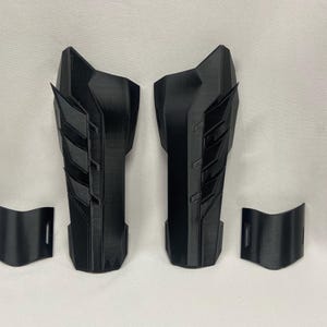 Puede incluir: Un conjunto de cuatro piezas impresas en 3D de color negro, dos piezas largas con un diseño geométrico y dos piezas más cortas. Las piezas son probablemente para un disfraz o cosplay.