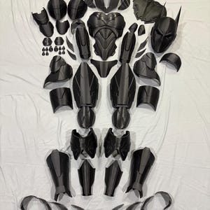 Könnte beinhalten: Eine Ansammlung schwarzer Kostümteile, möglicherweise für Cosplay, auf weißem Hintergrund angeordnet. Das Set umfasst einen Helm mit markanten Ohren, Brust- und Schulterpanzer, Bein- und Armschützer sowie weitere Elemente. Die Rüstung scheint aus einem starren Material zu bestehen.