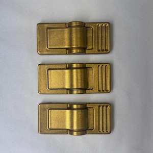 Può includere: Tre clip rettangolari in metallo dorato disposte verticalmente. Ogni clip presenta un meccanismo centrale incernierato con una superficie liscia e arrotondata e una sezione finale testurizzata con tre scanalature verticali parallele.