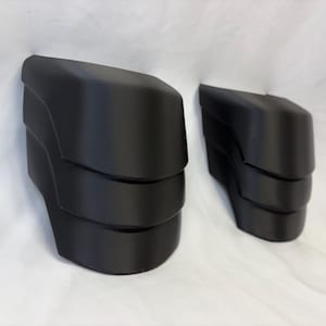 May include: Two black, angular automotive parts are shown against a white backdrop. The parts have a layered design, with a curved top and a series of horizontal bands. The objects appear to be made of a matte material.