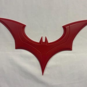May include: A red, flat, symmetrical emblem shaped like a stylised bat with pointed wings and a central head section. The design features recessed lines that follow the contours of the wings and body, creating a defined, layered appearance.