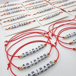 Pode incluir: Pulseiras de fio vermelho com contas de letras brancas que soletram "ARIRANG" e contas douradas. As pulseiras s&atilde;o exibidas em cart&otilde;es brancos, com v&aacute;rias pulseiras soltas em primeiro plano.