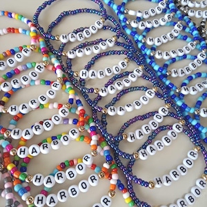 May include: A collection of colorful beaded bracelets. Each bracelet features the word "HARBOR" spelled out in white letter beads. The bracelets are in various colors, including blue, purple, and rainbow hues, with gold accents.