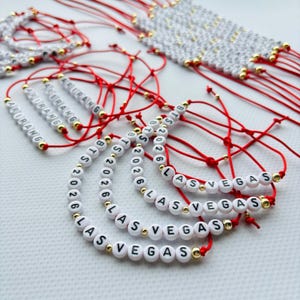 Pode incluir: Uma cole&ccedil;&atilde;o de pulseiras artesanais com contas de letras brancas que soletram "ARIRANG", "BTS 2026" e "LAS VEGAS". As pulseiras s&atilde;o amarradas em cord&otilde;es vermelhos com contas douradas. Algumas pulseiras s&atilde;o amarradas.