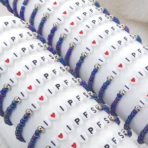 May include: A collection of beaded bracelets. Each bracelet features white letter beads spelling "IPFS" with a red heart symbol, interspersed with iridescent blue and purple beads. Silver-colored beads separate the sections.