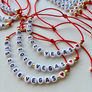 Pode incluir: Pulseiras de contas brancas com letras pretas que soletram "BTS 2026 VEGAS" e outro texto. As pulseiras s&atilde;o enfiadas em cord&otilde;es vermelhos com contas douradas e pequenas contas vermelhas em forma de cora&ccedil;&atilde;o. Em uma superf&iacute;cie branca.