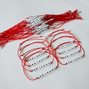 Pode incluir: Pulseiras de fio vermelho com contas brancas com letras que soletram "ARIRANG". V&aacute;rias pulseiras s&atilde;o vis&iacute;veis, algumas dispostas em um feixe, outras individualmente. As contas s&atilde;o brancas com letras pretas.