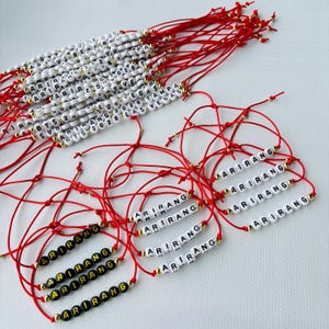 Pode incluir: Uma cole&ccedil;&atilde;o de pulseiras artesanais com cord&otilde;es vermelhos e contas de letras brancas ou pretas que soletram "ARIRANG". Algumas pulseiras t&ecirc;m texto adicional como "BTS 2026 LAS VEGAS". Contas douradas acentuam o design.