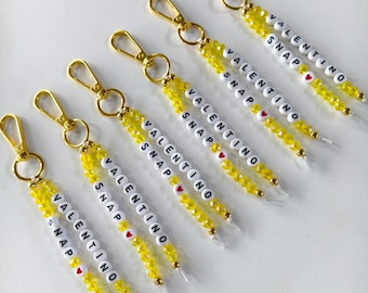 May include: Five gold-toned lobster clasp keychains featuring strands of yellow faceted beads and white letter beads spelling out VALENTINO SNAP with a small red heart bead. Each keychain is identical in design, color, and arrangement of beads.