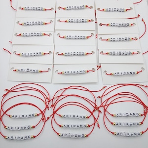 Pode incluir: Pulseiras de fio vermelho com a palavra "ARIRANG" escrita em contas de letras brancas. As pulseiras s&atilde;o exibidas em cart&otilde;es brancos e tamb&eacute;m separadamente. Contas douradas acentuam o design.