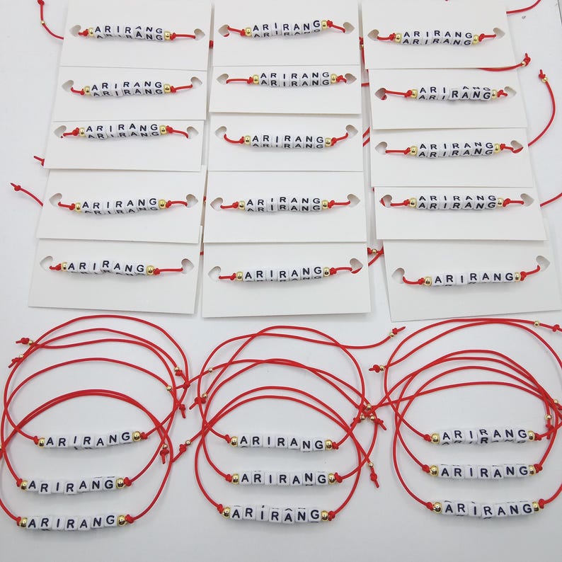 Pode incluir: Pulseiras de fio vermelho com a palavra "ARIRANG" escrita em contas de letras brancas. As pulseiras s&atilde;o exibidas em cart&otilde;es brancos e tamb&eacute;m separadamente. Contas douradas acentuam o design.