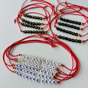 Pode incluir: V&aacute;rias pulseiras de fio vermelho. Algumas soletrando "ARIRANG" com contas pretas e douradas, outras t&ecirc;m "ARIRANG 2026" com contas brancas e douradas. As pulseiras est&atilde;o em uma superf&iacute;cie branca.