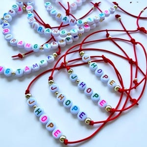 Pode incluir: V&aacute;rias pulseiras artesanais com contas de letras brancas que soletram nomes e a palavra "HOPE". As pulseiras s&atilde;o amarradas em cord&otilde;es vermelhos com detalhes dourados. As letras s&atilde;o de v&aacute;rias cores.