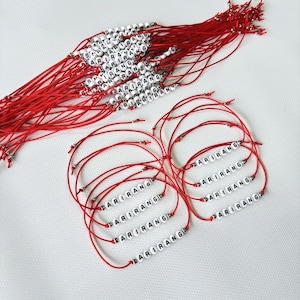 Pode incluir: Pulseiras de fio vermelho com contas de letras brancas que soletram "ARIRANG". As pulseiras s&atilde;o amarradas com n&oacute;s e t&ecirc;m um design simples. Um monte de pulseiras semelhantes &eacute; vis&iacute;vel no fundo.