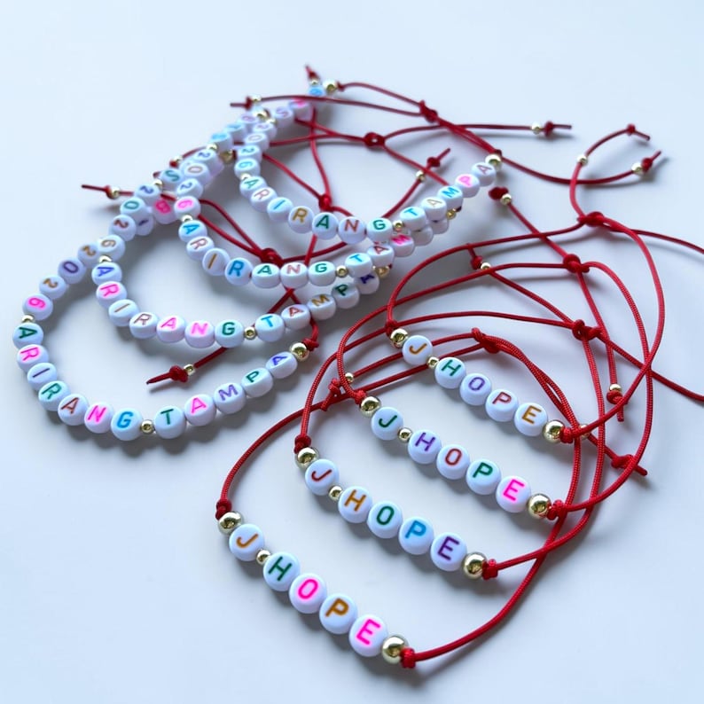Pode incluir: V&aacute;rias pulseiras artesanais com contas de letras brancas que soletram nomes e a palavra "HOPE". As pulseiras t&ecirc;m cord&otilde;es vermelhos e detalhes dourados. As contas de letras s&atilde;o de v&aacute;rias cores.