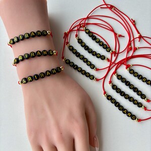 Pode incluir: Pulseiras de contas pretas com a palavra "ARIRANG" em letras douradas. As pulseiras s&atilde;o enfiadas em cord&otilde;es vermelhos com detalhes dourados. V&aacute;rias pulseiras s&atilde;o exibidas em um pulso e separadamente.