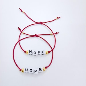Pode incluir: Duas pulseiras de fio vermelho com a palavra "HOPE" escrita com contas de letras brancas. As pulseiras t&ecirc;m contas douradas e s&atilde;o apresentadas em um fundo branco. As pulseiras s&atilde;o ajust&aacute;veis.