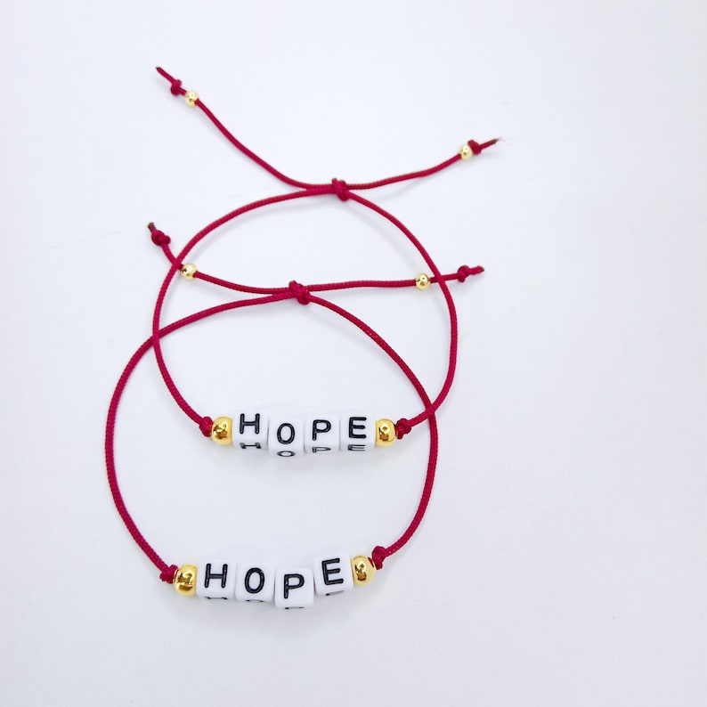 Pode incluir: Duas pulseiras de fio vermelho com a palavra "HOPE" escrita com contas de letras brancas. As pulseiras t&ecirc;m contas douradas e s&atilde;o apresentadas em um fundo branco. As pulseiras s&atilde;o ajust&aacute;veis.