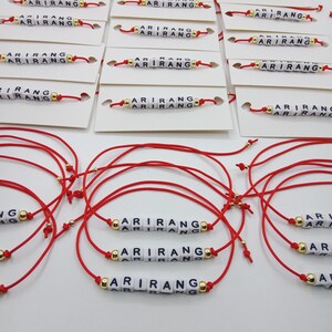 Pode incluir: Pulseiras de cord&atilde;o vermelho com a palavra "ARIRANG" escrita em contas de letras brancas. As pulseiras t&ecirc;m contas douradas e s&atilde;o exibidas em cart&otilde;es brancos.