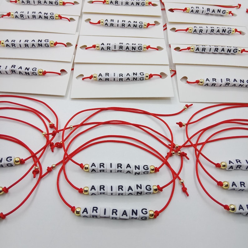 Pode incluir: Pulseiras de cord&atilde;o vermelho com a palavra "ARIRANG" escrita em contas de letras brancas. As pulseiras t&ecirc;m contas douradas e s&atilde;o exibidas em cart&otilde;es brancos.