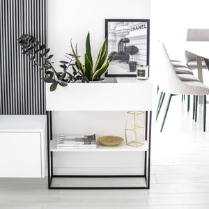 Könnte beinhalten: Weißes Regal mit schwarzem Metallrahmen, zwei Regalböden und einem Pflanztopf. Das obere Regal hat ein goldenes Dekotablett, einen Kerzenhalter aus Glas und eine weiße Kerze. Der Pflanztopf hat eine grüne Pflanze und einen Schwarzweißdruck eines Chanel-Ladens mit dem Text "CHANEL" darauf.