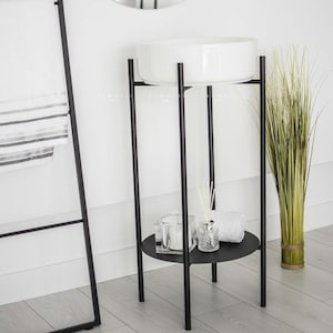 Op de afbeelding: Een witte keramische wastafel op een zwarte metalen standaard met een rond plankje. De standaard heeft vier poten en een rond bovenste plankje. Op het plankje staan een klein glazen potje, een flesje met diffuserstokjes en een witte handdoek.