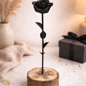 Puede incluir: Una escultura de rosa de metal negro con pétalos y hojas detallados. La rosa está montada sobre un tallo oscuro y delgado y colocada en una base de rodaja de madera natural. Una idea de regalo decorativa.