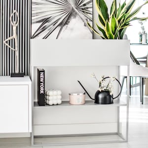 Pode incluir: Uma estante branca com duas prateleiras, uma impressão em preto e branco de uma palmeira, um livro preto intitulado "NEW YORK", uma vela branca, um regador preto com flores brancas e uma planta verde em um vaso branco.