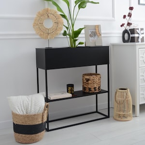 Pode incluir: Um suporte para plantas de metal preto com duas prateleiras. A prateleira superior tem um vaso com uma planta verde e uma cesta tecida. A prateleira inferior tem um livro, uma vela e outra cesta tecida. O suporte está em frente a uma parede branca com lambrim.