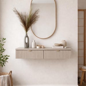 Op de afbeelding: Een interieur in neutrale tinten met een aan de muur gemonteerde kast met geribbelde details, een ovaalvormige spiegel met gouden lijst en decoratieve items. Een gevlochten mand met een plant en een rond jute vloerkleed voegen textuur toe.
