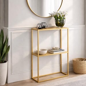 Op de afbeelding: Een gouden metalen console met twee planken, met een witte kom, boeken en decoratieve items. Een ronde gouden spiegel hangt erboven. Een geweven mand en een plant in pot staan ook in de kamer. Een grijze vloerkleed ligt op de vloer.
