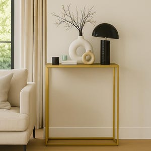 Puede incluir: Un salón moderno con una consola dorada. La mesa exhibe una lámpara negra, jarrones blancos y beige, y objetos decorativos. Un sofá y una alfombra beige también están presentes, creando una estética minimalista.