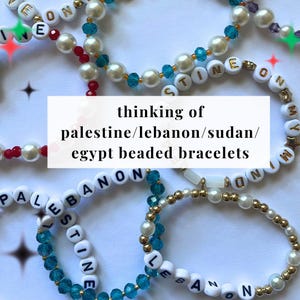 May include: Close-up of four beaded bracelets with the words "Palestine", "Lebanon", "Sudan", and "Egypt" spelled out in white letters. The bracelets are made with white and blue beads.