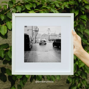 Puede incluir: Una fotografía en blanco y negro de una calle de la ciudad con un coche pequeño conduciendo por el medio de la calle. La calle está mojada por la lluvia y hay edificios a ambos lados de la calle.