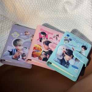 May include: Three colourful faux leather journals with snap closures. The journals are lavender, pink, and light blue. Each journal features a different name and images of a person. The lavender journal says "Xavier", the pink journal says "Rayael", and the light blue journal says "Zayne".