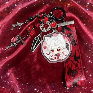 May include: A gothic-themed keychain with a black ring, featuring charms like a coffin, a sword with a rose, and a pendant with a cartoon character. Red and black beads and a red ribbon with black designs complete the design.