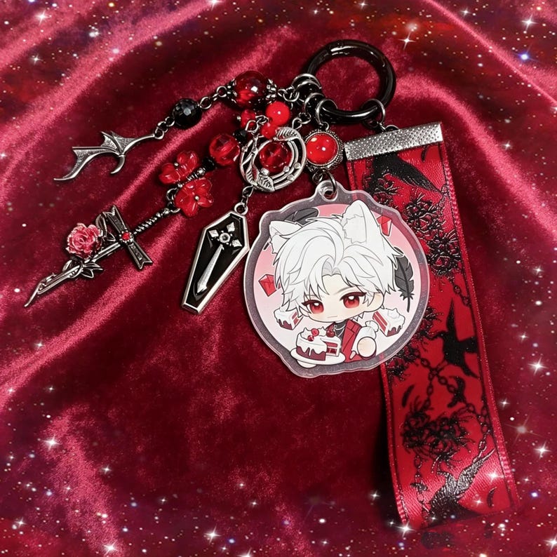May include: A gothic-themed keychain with a black ring, featuring charms like a coffin, a sword with a rose, and a pendant with a cartoon character. Red and black beads and a red ribbon with black designs complete the design.